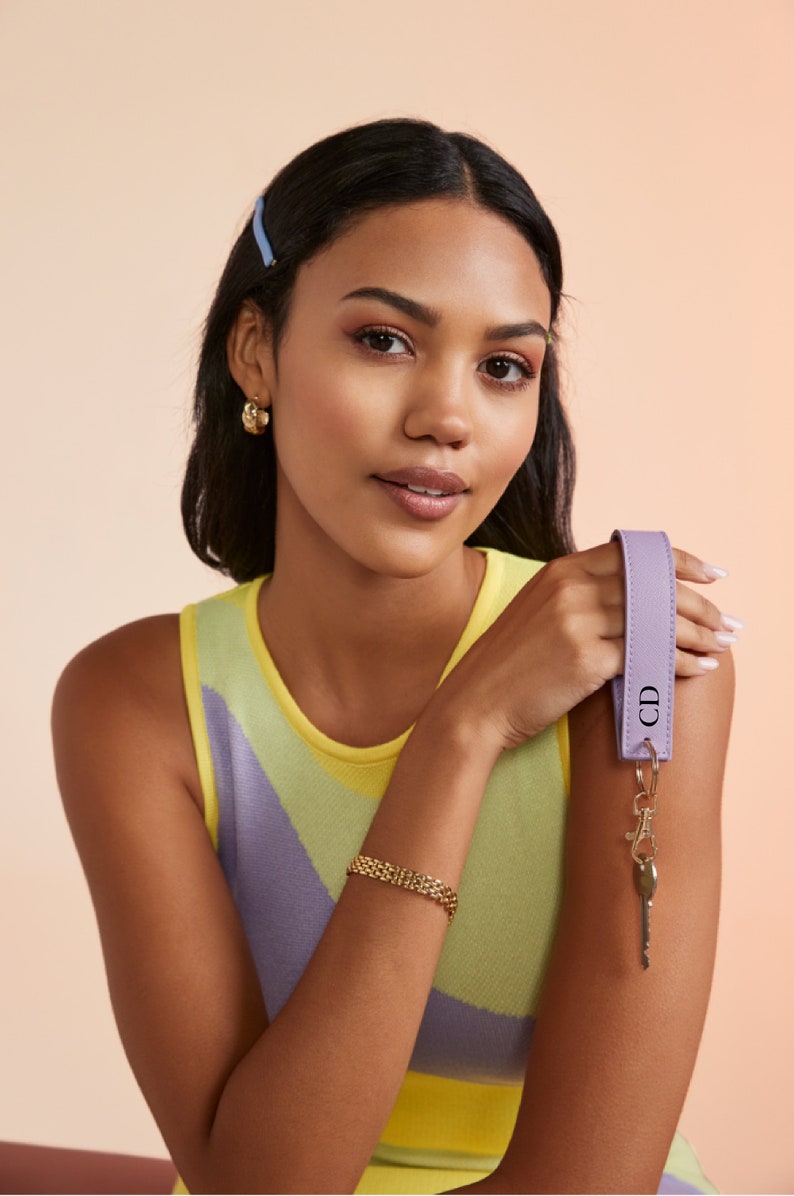K&ouml;nnte beinhalten: Eine Frau tr&auml;gt ein gelbes und violettes, &auml;rmelloses Oberteil mit einem goldenen Gliederarmband. Sie h&auml;lt einen lilafarbenen Leder-Schl&uuml;sselanh&auml;nger mit den Initialen "CD" in der Hand.