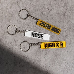 May include: Three keychains with silver key rings and chains. The keychains are rectangular and made of clear acrylic with yellow and white backgrounds. The text on the keychains reads "25th Aug", "Rose", and "KIGH X R".