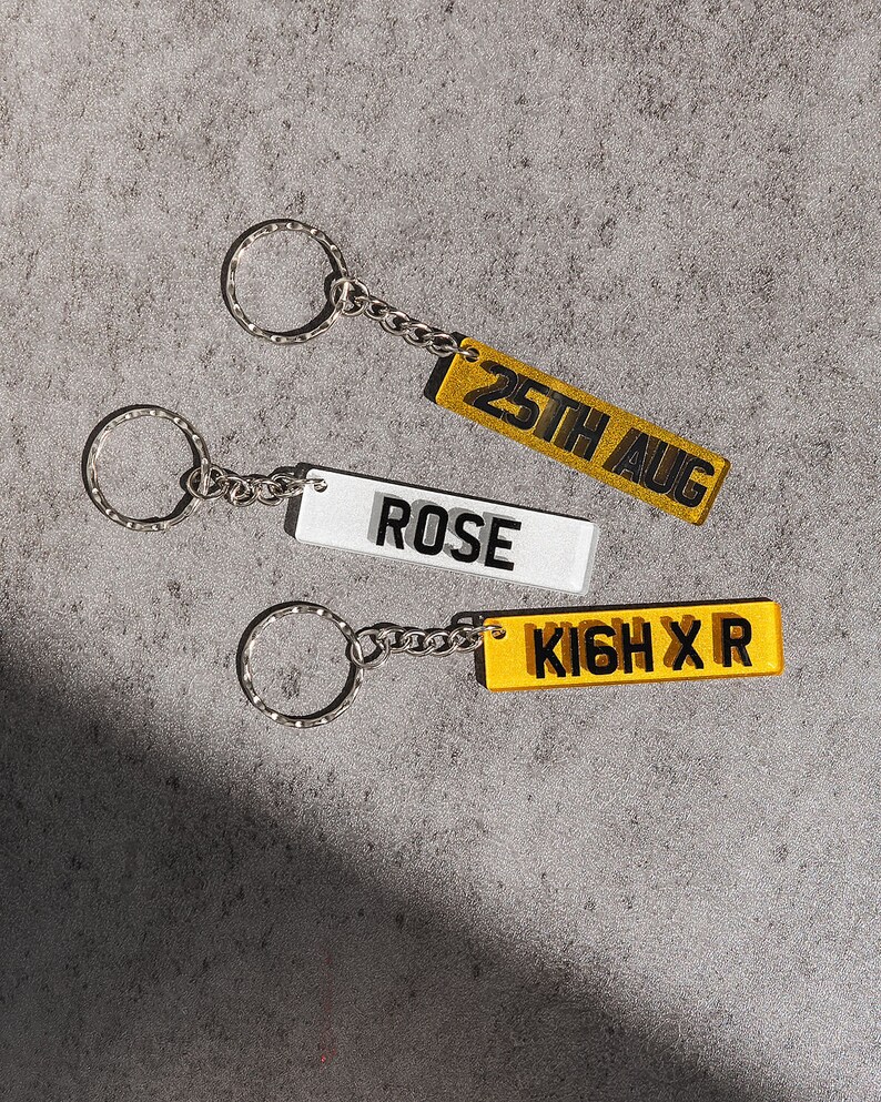 May include: Three keychains with silver key rings and chains. The keychains are rectangular and made of clear acrylic with yellow and white backgrounds. The text on the keychains reads "25th Aug", "Rose", and "KIGH X R".