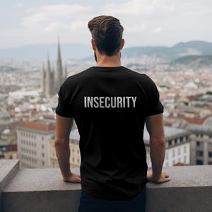 Może przedstawiać: Czarny t-shirt z białym napisem "INSECURITY" na plecach. Osoba stoi na gzymsie z widokiem na miasto z wysokimi budynkami i kościołem.