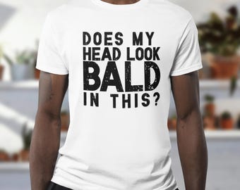 May include: White t-shirt with the text "DOES MY HEAD LOOK BALD IN THIS?" in a bold, distressed black font. The shirt is a crew neck style and appears to be made of a soft material. The background is blurred, suggesting an indoor setting.