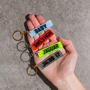 May include: Four colorful keychains with different names and words on them. The keychains are attached to a set of gold and silver key rings. The keychains are held in a person's hand.