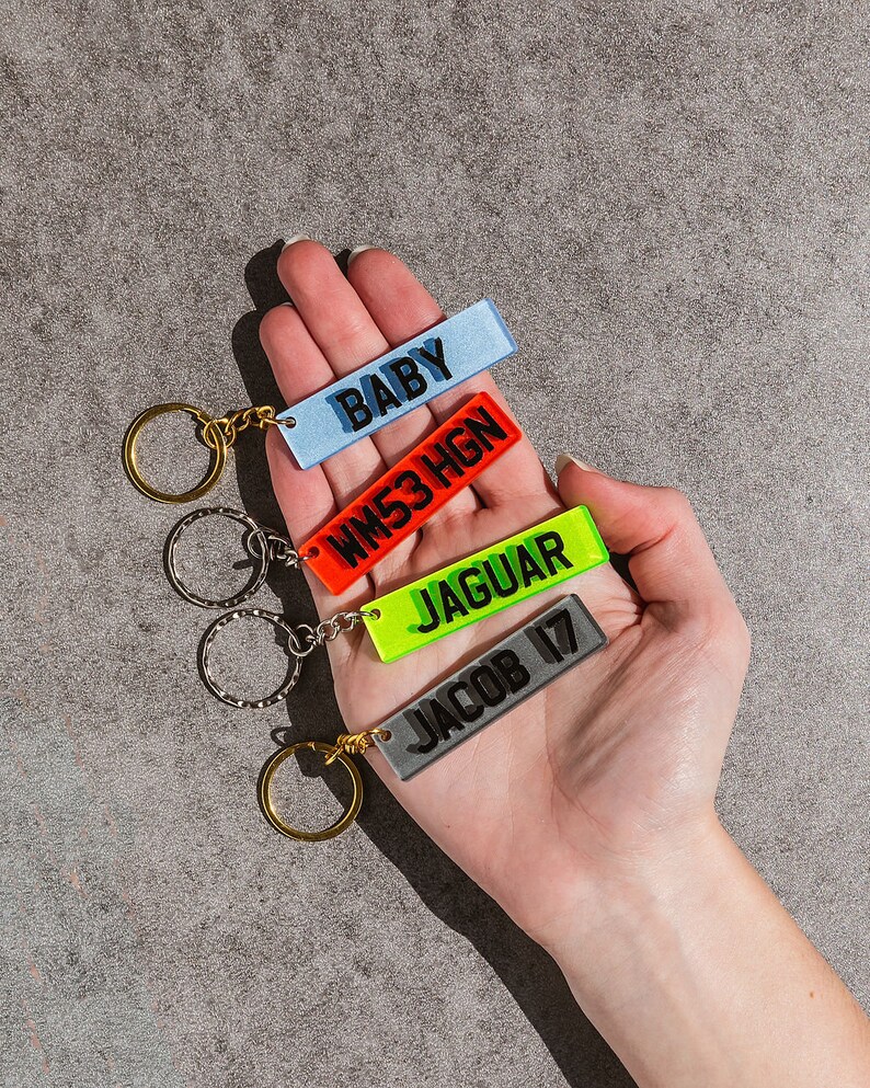 May include: Four colorful keychains with different names and words on them. The keychains are attached to a set of gold and silver key rings. The keychains are held in a person's hand.