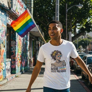 Könnte beinhalten: Weißes T-Shirt mit einer Grafik einer Frau mit Regenbogen-Sonnenbrille und Blumen. Der Text "MORE PRIDE LESS PREJUDICE" steht darunter. Im Hintergrund ist eine Regenbogenflagge zu sehen.