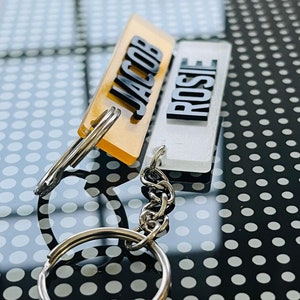 May include: Two acrylic keychains with the names "JACOB" and "ROSIE" on them. The keychain with the name "JACOB" is yellow and the keychain with the name "ROSIE" is white. The keychains are attached to a silver chain.
