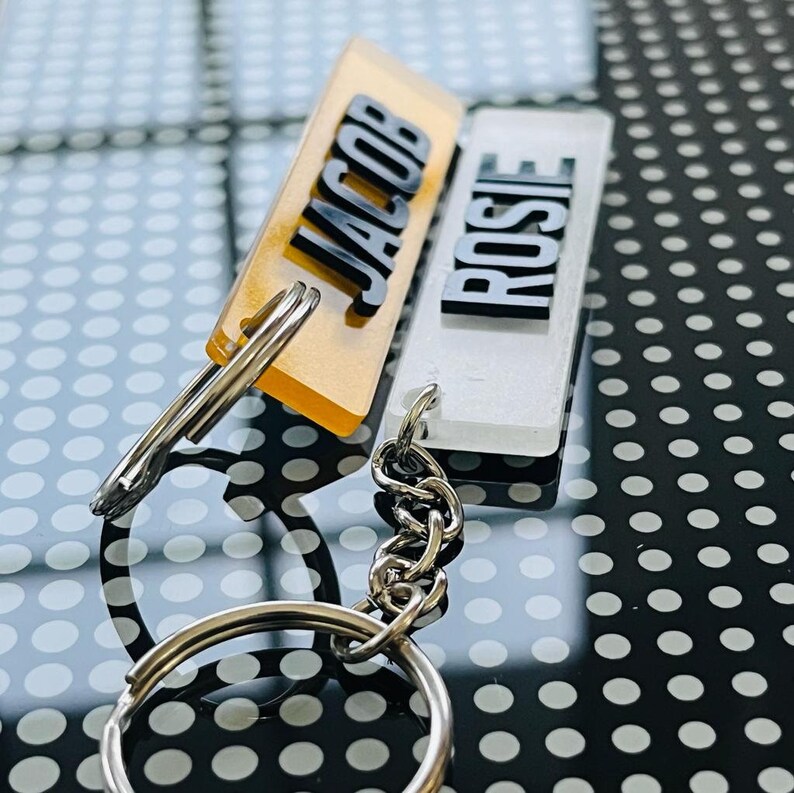 May include: Two acrylic keychains with the names "JACOB" and "ROSIE" on them. The keychain with the name "JACOB" is yellow and the keychain with the name "ROSIE" is white. The keychains are attached to a silver chain.
