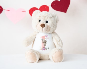 May include: A cream-colored teddy bear with a white t-shirt featuring a pink number 1, a cartoon hippo holding a star balloon, and the name "Freya". The bear is decorated with floral accents and is set against a backdrop of heart-shaped decorations.