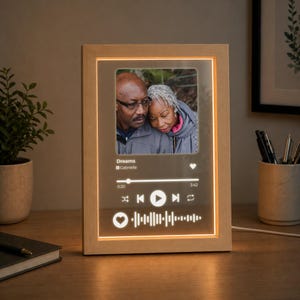 May include: A rectangular light-up frame with a light wood finish containing a transparent panel. The panel displays a photograph of two people, the song title Dreams by Gabrielle, a playback progress bar, media controls, and a sound wave graphic.