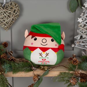 May include: A plush elf toy with a green hat and red and green clothing. The elf has a smiling face and the text "Theo's 1st Christmas 2024" is embroidered on the front.