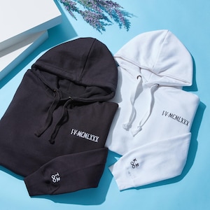 May include: Two hooded sweatshirts, one black and one white, are displayed on a light blue surface. Each sweatshirt has the text "I-V-MCMLXXX" embroidered in white. The black sweatshirt has the initials "I.R" and the number "88" embroidered on the sleeve.
