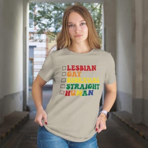 May include: A beige t-shirt with the words "LESBIAN, GAY, BISEXUAL, STRAIGHT, HUMAN" in rainbow colors. The word "HUMAN" is checked. The shirt is short-sleeved.