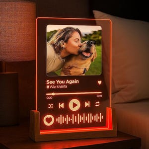 Puede incluir: Panel acrílico rectangular iluminado con foto de una persona besando a un perro. El panel muestra el título de la canción, el artista y los controles de reproducción. La base es de madera, los bordes brillan en naranja.