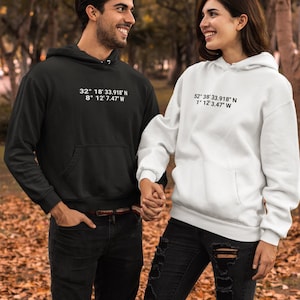 Peut inclure: Un sweat-shirt à capuche noir avec le texte blanc 32° 18' 33.918" N 8° 12' 7.47" W et un sweat-shirt à capuche blanc avec le texte noir 52° 38' 33.918" N 1° 12' 3.47" W, tous deux dotés de manches longues, d'une poche kangourou et d'une capuche.