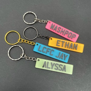 Op de afbeelding: Vier kleurrijke sleutelhangers met zwarte tekst. De sleutelhangers zijn roze, geel, blauw en groen. De tekst op de sleutelhangers is "Washpop", "Ethan", "LCFC Jay" en "Alyssa".