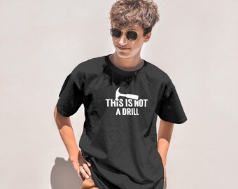 Op de afbeelding: Zwart t-shirt met de witte tekst "THIS IS NOT A DRILL" en een hamerafbeelding. De persoon draagt een zonnebril en een bruine broek.