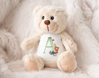 May include: A plush beige teddy bear with a white shirt featuring a green letter "A", a red flower, and a small brown bear illustration. The shirt also includes the name "Asher" and the date 06.03.23.