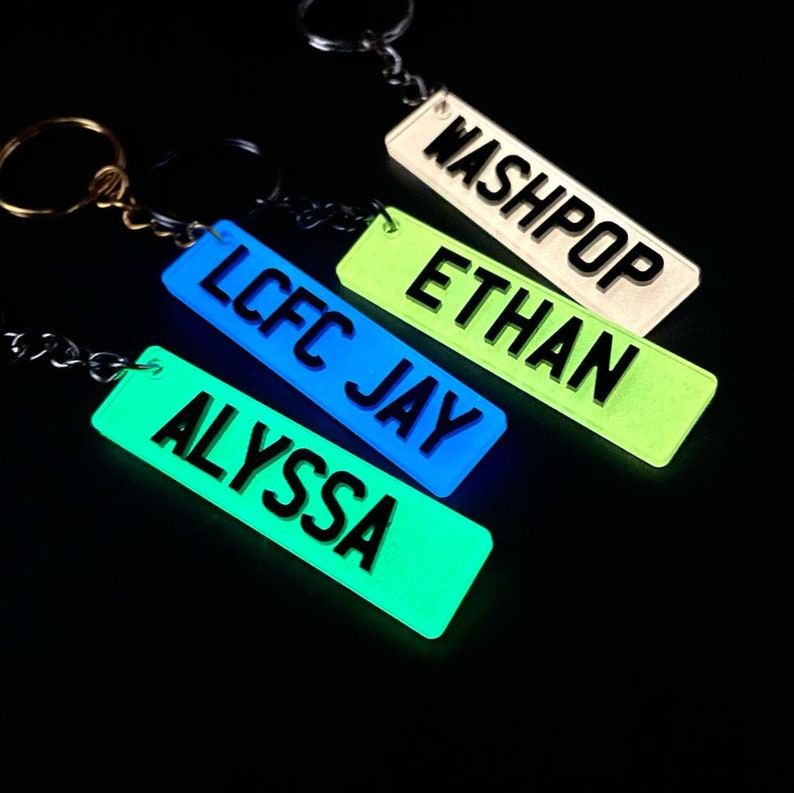 Op de afbeelding: Vier glow-in-the-dark sleutelhangers met verschillende namen: Washpop, Ethan, LCFC Jay en Alyssa. De sleutelhangers zijn allemaal in verschillende kleuren: wit, groen, blauw en groen.