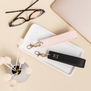 K&ouml;nnte beinhalten: Zwei personalisierte Leder-Schl&uuml;sselanh&auml;nger mit goldenen Buchstaben. Der Schl&uuml;sselanh&auml;nger links ist rosa mit den Initialen "OG" und der Schl&uuml;sselanh&auml;nger rechts ist schwarz mit den Initialen "EJ".