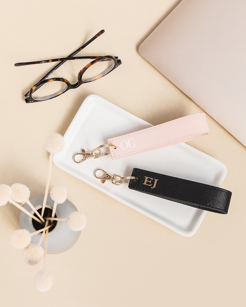 K&ouml;nnte beinhalten: Zwei personalisierte Leder-Schl&uuml;sselanh&auml;nger mit goldenen Buchstaben. Der Schl&uuml;sselanh&auml;nger links ist rosa mit den Initialen "OG" und der Schl&uuml;sselanh&auml;nger rechts ist schwarz mit den Initialen "EJ".
