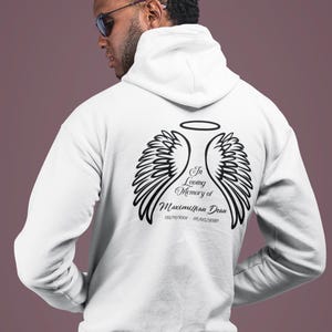 May include: A white hooded sweatshirt featuring a black graphic on the back. The design includes two angel wings, a halo, and the text In Loving Memory of Maximilian Dean with the dates 02/11/1991 - 07/05/2020 printed in a script font.
