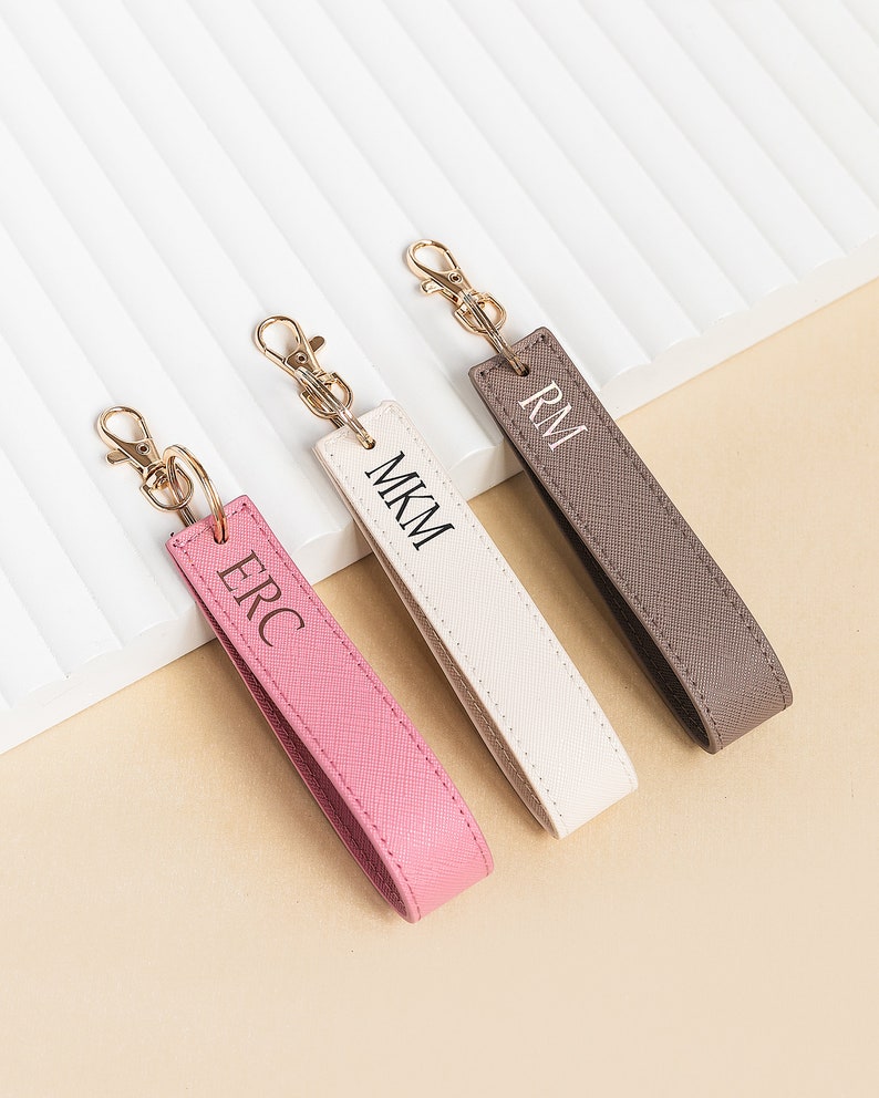 K&ouml;nnte beinhalten: Drei personalisierte Leder-Schl&uuml;sselanh&auml;nger mit goldenen Schl&uuml;sselringen. Die Schl&uuml;sselanh&auml;nger sind in rosa, wei&szlig; und braun mit den Initialen "ERC", "MKM" und "RM".