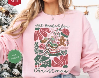 May include: Light pink sweatshirt with the text "All Booked for Christmas" and a graphic of a Christmas tree made of books, surrounded by festive elements like candy canes, ornaments, and a gingerbread man.