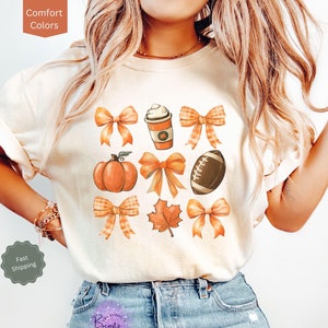 May include: A white t-shirt with a fall-themed design featuring a pumpkin, football, coffee cup, maple leaf, and bows in orange and brown.