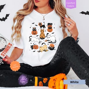 May include: White t-shirt with a Halloween cat design. The design features multiple cats in various poses, wearing witch hats, holding pumpkins, and surrounded by bats and stars. The text "Comfort Colors" is visible in the upper right corner of the image.