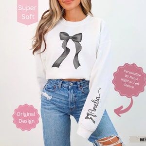 May include: White sweatshirt with a large gray bow graphic on the front. The sleeve features the name "Amelia" in a script font. The text "Super Soft", "Original Design", and "Personalize W/ Name Right or Left Sleeve" are also visible.