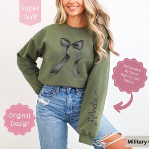 May include: Military green sweatshirt with a black bow graphic. The long-sleeved top features the text "Amelia" on the right sleeve. The design is described as original and super soft. Personalization with a name is available on the right or left sleeve.