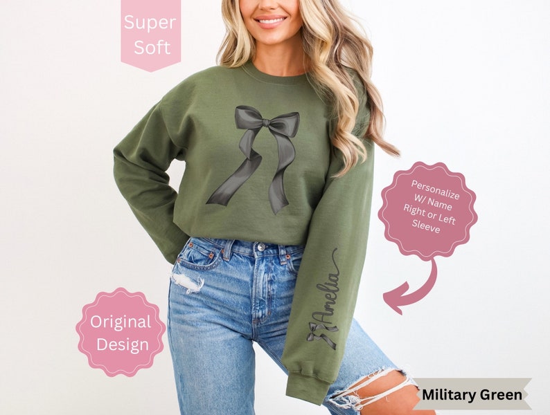 May include: Military green sweatshirt with a black bow graphic. The long-sleeved top features the text "Amelia" on the right sleeve. The design is described as original and super soft. Personalization with a name is available on the right or left sleeve.