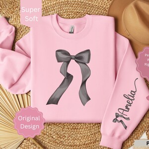 May include: A pink sweatshirt with a large gray bow graphic on the front and a smaller bow with the name "Amelia" on the sleeve. The words "Super Soft", "Original Design", and "Pink" are also visible.