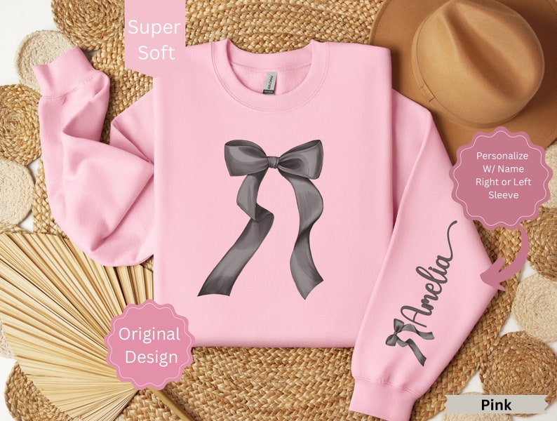 May include: A pink sweatshirt with a large gray bow graphic on the front and a smaller bow with the name "Amelia" on the sleeve. The words "Super Soft", "Original Design", and "Pink" are also visible.
