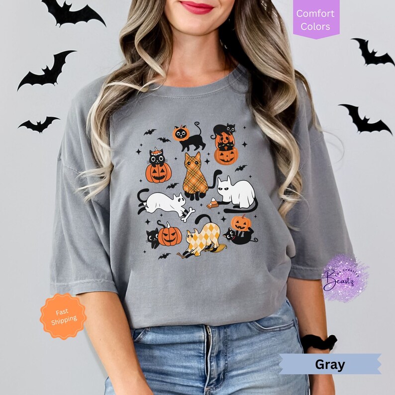 May include: Gray t-shirt with a Halloween design featuring black cats and pumpkins. The cats are wearing hats and some are holding bones. The text "Comfort Colors" is in the upper right corner of the image.