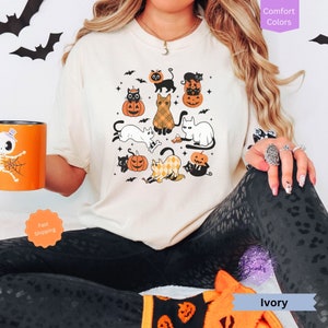 May include: A white t-shirt with a graphic of black and white cats wearing pumpkin hats and holding pumpkins. The graphic also includes bats and stars. The shirt is on a model wearing black leggings with a leopard print.