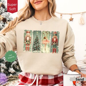 May include: A beige sweatshirt with a Christmas-themed design featuring a ballerina, two nutcrackers, and a Christmas tree. The design is in a vintage style with a green, red, and gold color scheme. The sweatshirt has a crew neck and long sleeves.