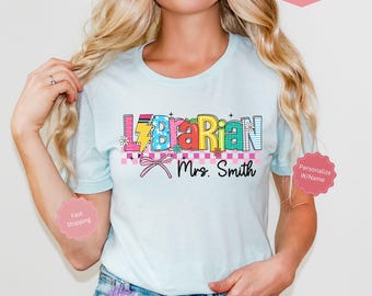 Op de afbeelding: Een lichtblauw T-shirt met een kleurrijke afbeelding waarop "Librarian" staat met een bliksem en bloemen. De tekst "Mrs. Smith" staat onder de afbeelding.