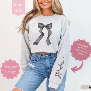 May include: Ash-colored sweatshirt featuring a gray bow design on the front. The right sleeve is personalized with the name "Amelia" in a script font. The sweatshirt is described as "Super Soft" and an "Original Design".