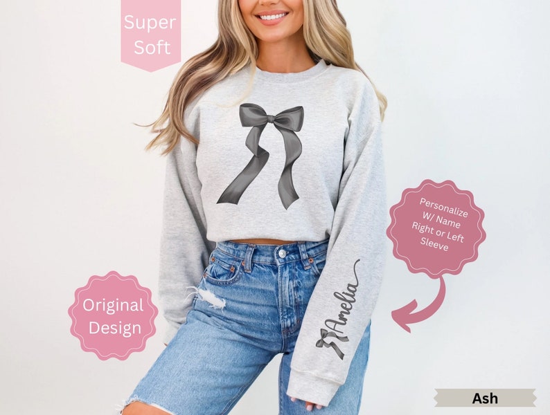 May include: Ash-colored sweatshirt featuring a gray bow design on the front. The right sleeve is personalized with the name "Amelia" in a script font. The sweatshirt is described as "Super Soft" and an "Original Design".