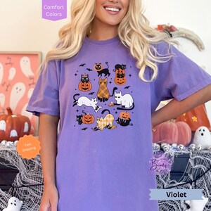 May include: Purple t-shirt with a Halloween design featuring black and white cats, pumpkins, and bats. The text "Violet" is visible at the bottom of the image.