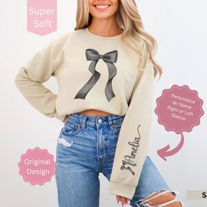 May include: Sand-colored sweatshirt featuring a large gray bow design on the front and the name "Amelia" in script on the sleeve. The sweatshirt is labeled "Super Soft" and "Original Design."