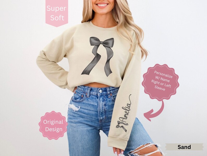 May include: Sand-colored sweatshirt featuring a large gray bow design on the front and the name "Amelia" in script on the sleeve. The sweatshirt is labeled "Super Soft" and "Original Design."