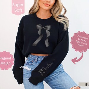 May include: Black sweatshirt featuring a gray bow design. The sleeve is personalized with a name in cursive. The text "Super Soft" and "Original Design" are also visible. The sweatshirt is black.