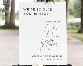 May include: A white wedding welcome sign on a black easel. The sign reads "WE'RE SO GLAD YOU'RE HERE." and "THE WEDDING OF Julia AND Matthew." The date is October 10, 20XX. White flowers and greenery decorate the top and bottom.