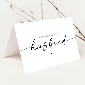 May include: A white folded card with the text "I can't wait to be your husband" in a stylish script font. A small heart is below the word "husband". The card is on a white surface with a twig in the background.
