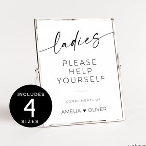 May include: A white sign with black text that reads "ladies please help yourself" and "compliments of Amelia Oliver". The sign is on a silver easel and has a black circle with white text that reads "includes 4 sizes".