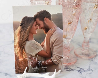 Puede incluir: Una tarjeta blanca con una foto de una pareja abrazándose, el texto "Table One" en cursiva, y los nombres "Amelia + Oliver" y la fecha "08.17.20XX". La tarjeta está apoyada en una asta de ciervo blanca.