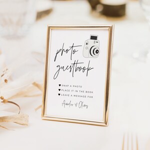 May include: A white sign with a gold frame and a black and white illustration of a camera. The sign says "photo guestbook" and includes instructions for guests to snap a photo, place it in the book, and leave a message for "Amdir & Oliver".