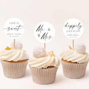 Pode incluir: Tr&ecirc;s cupcakes brancos com cobertura branca e granulado dourado. Cada cupcake tem um topo redondo de papel branco com texto preto. Os topos dizem "Love is sweet Amelia + Oliver 08.20.20XX", "Mr. & Mrs." e "Happily Ever After! Amelia + Oliver 08.20.20XX".