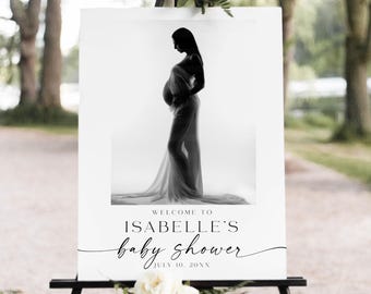 May include: A black and white baby shower sign featuring a silhouette of a pregnant person draped in fabric. The sign reads "Welcome to Isabelle's baby shower" with the date July 10, 20XX. The sign is displayed on a black easel.